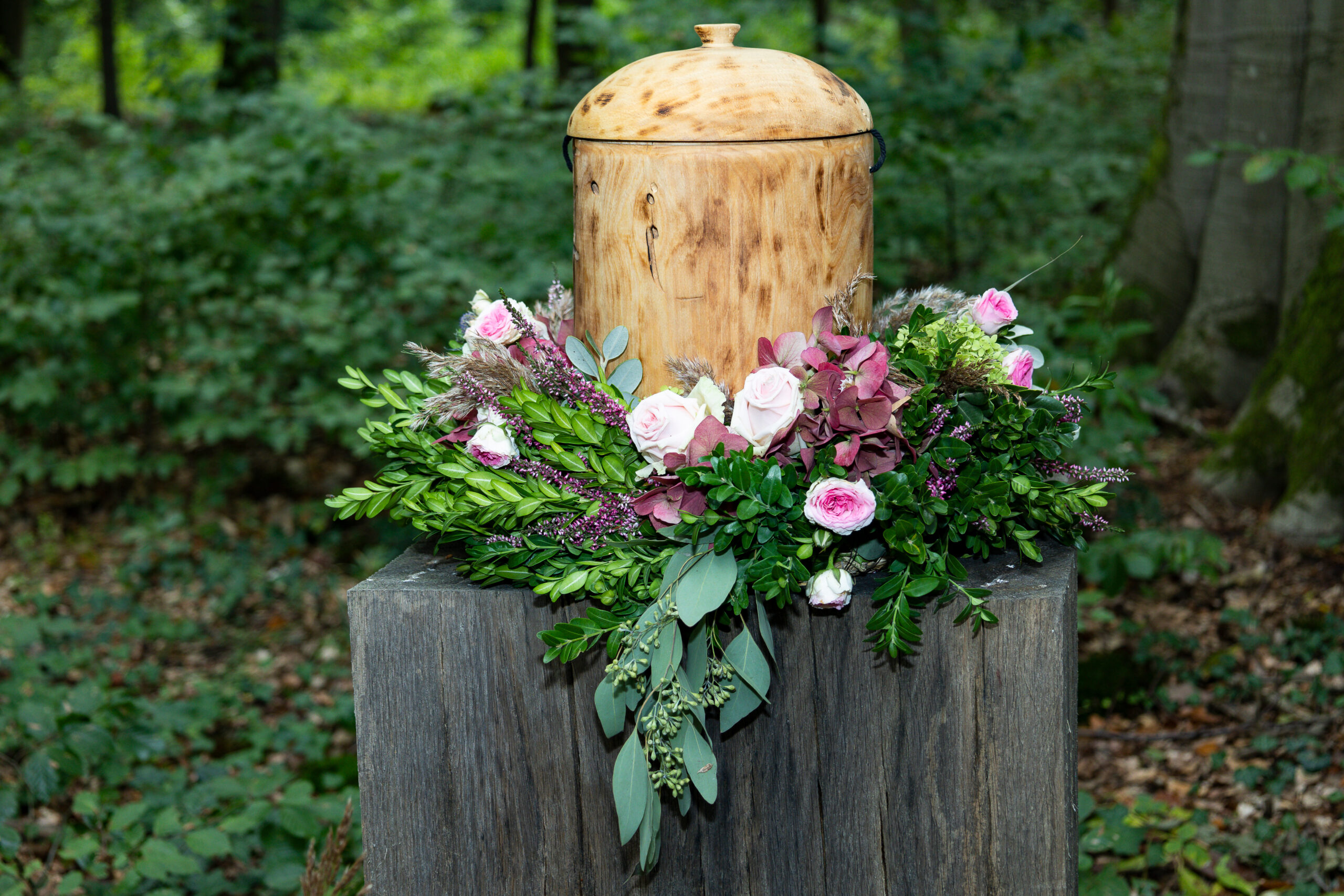 A funeral urn of ashes surrounded by an arrangement of pink flowers on a tree stump in an outdoor setting